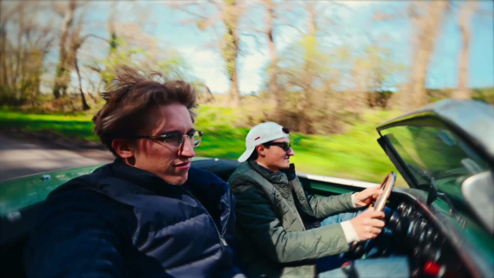 Two people driving a classic green convertible down a tree-lined road
